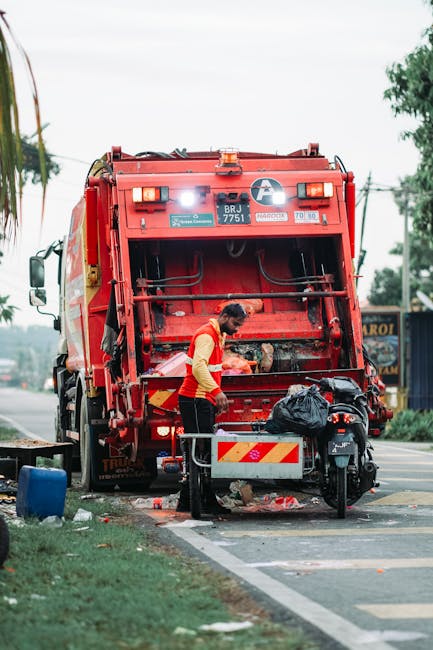 A municipal worker dressed in a high-visibility yellow and red vest is operating a large, red waste collection truck on a street. The truck, part of an independent rubbish removal service, has its rear compactor door open, revealing internal mechanisms used for compacting refuse. The worker is in the process of loading waste materials, with a black plastic rubbish bag placed nearby on a small wheeled trolley. The scene includes details of the roadside environment, such as a blue plastic container on the grass verge, a motorcycle parked adjacent to the worker, and a partly visible signboard in the background. Overhead, power lines and trees are visible against a cloudy sky, indicating a typical urban or suburban setting. The truck's bright red finish and safety reflective markings contrast with the surrounding environment, emphasizing its utility for private waste handling and rubbish removal services that support local disposal needs outside of council collections.