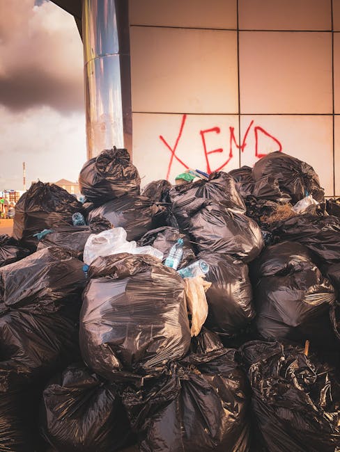 A large pile of black rubbish bags, some partially torn or bulging, is stacked against a modern building with a reflective tiled wall. The bags appear to contain waste materials and are situated outdoors, likely in an urban setting. Several clear plastic bottles are visible among the bags, indicating plastic waste disposal. Behind the pile, the building has a smooth surface with a grid of beige or light brown tiles and prominent red graffiti spelling 'XEND' on the wall. The scene is illuminated by natural daylight, with a cloudy sky visible in the background, creating a somewhat overcast atmosphere. The overall setting suggests an area awaiting collection or disposal, possibly related to private waste management services such as those offered by Rubbish Removal Bermondsey, highlighting the need for efficient rubbish removal or on-site clearance solutions in the borough.