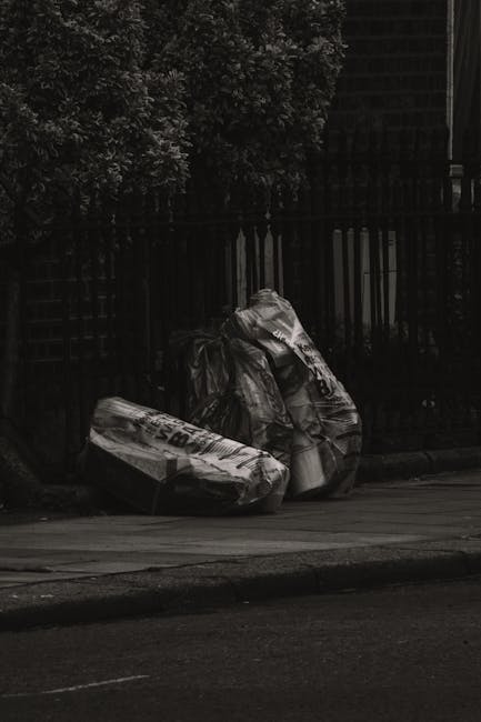 A large, dirty, beige sack made of fabric, visibly stained and worn, is placed on the pavement next to a metal gate in an urban residential street. The sack appears to be filled with waste or discarded items, with some loose debris and a plastic bag protruding from its top. The surrounding environment features a narrow, uneven asphalt road flanked by old, weathered buildings with graffiti and iron bars on windows. In the background, laundry is hanging on clotheslines strung between buildings, and there are scattered pieces of trash and debris along the street, indicating a lack of immediate rubbish collection. The lighting is subdued, suggesting overcast weather or late afternoon conditions. This scene reflects a typical urban setting where independent waste disposal methods, such as placing refuse in sacks beside buildings, are used, aligning with the context of private rubbish removal services provided by companies like Rubbish Removal Bermondsey.