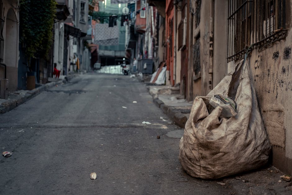 A narrow urban alleyway cluttered with various types of rubbish and waste materials. In the foreground, a large, grey, weathered bin with visible creases and dirt is positioned centrally, facing away from the camera. Behind the bin, stacks of cardboard boxes, some damaged and disorganized, are piled up, creating a makeshift wall of waste. The ground surface is dark and grimy, with patches of dirt and small debris scattered around. On the left side, a black trash can lies on its side, partially obscured by the bin. To the right, the alley is bordered by a grey building with metal bars on the windows, and graffiti is visible along the lower part of the wall. The background shows a continuation of the alley with more waste piled against a wooden barrier, topped by leafless trees and the side of an older brick building, suggesting an environment in need of private or independent rubbish clearance services. The scene appears dimly lit, evoking a gritty, urban atmosphere aligned with rubbish removal and waste collection activities overseen by companies like Rubbish Removal Bermondsey.
