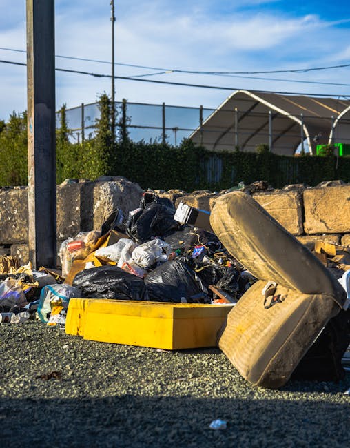 A close-up view of a pile of mixed domestic rubbish scattered on a gravel surface, including black plastic bin bags, a yellow plastic container, an old beige upholstered armchair with visible fabric texture and stains, and fragments of cardboard and plastic. The rubbish is piled against a low stone wall made of uneven, weathered stones, with some debris spilling onto the surrounding area. In the background, there are wooden utility poles with overhead electrical wires, a row of green shrubbery, and a large white canopy structure with a curved roof, indicating an outdoor setting near a storage or work yard. The scene is lit by daylight, capturing the textures and colors of discarded waste materials, suggesting a waste collection or clearance operation, potentially by a private rubbish removal service like Rubbish Removal Bermondsey.